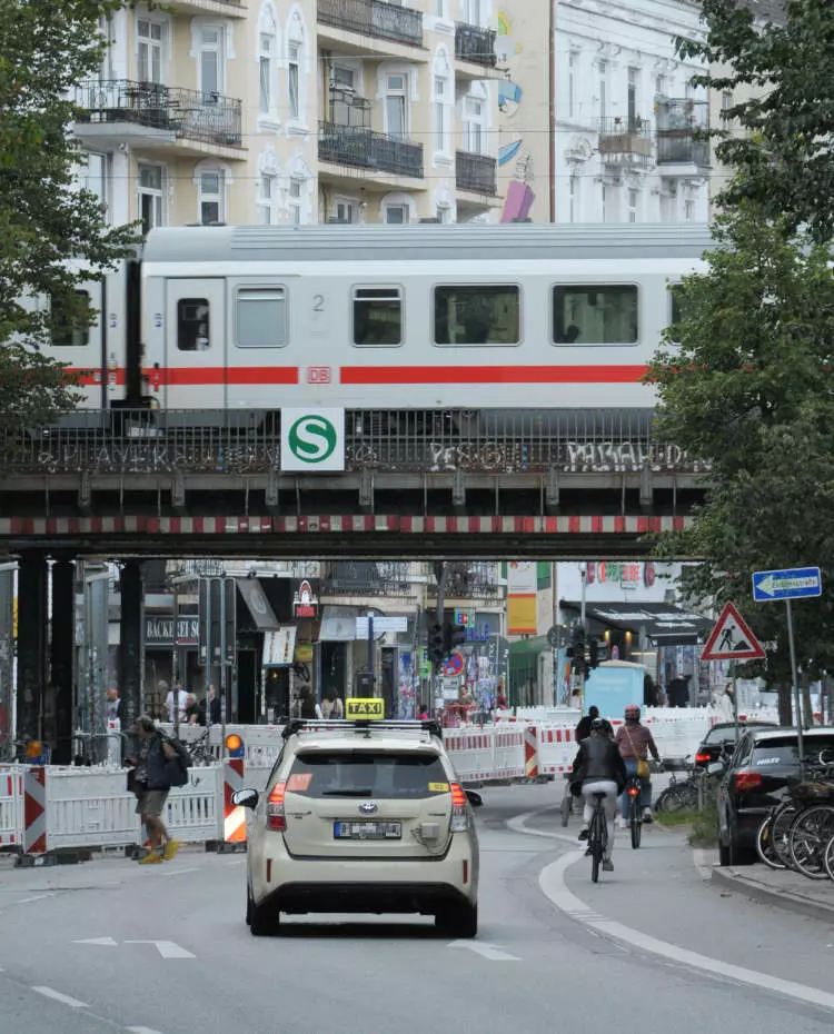Vorlage_Hochkant Ein Taxi fährt unter einer S-Bahn-Brücke durch, über die ein ICE fährt. Neben dem Taxi Fahrräder und Fußgänger. Foto: Unsplash|Riiana Izzietovo