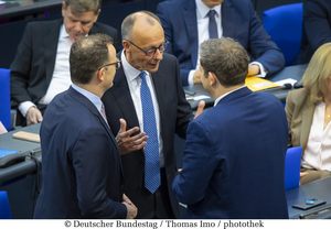 Friedrich Merz in der Mitte vom Bild im Gespräch mit Jens Spahn und Lars Klingbeil, die beide der Kamera den Rücken zuwenden. Alle stehen im Plenarsaal.