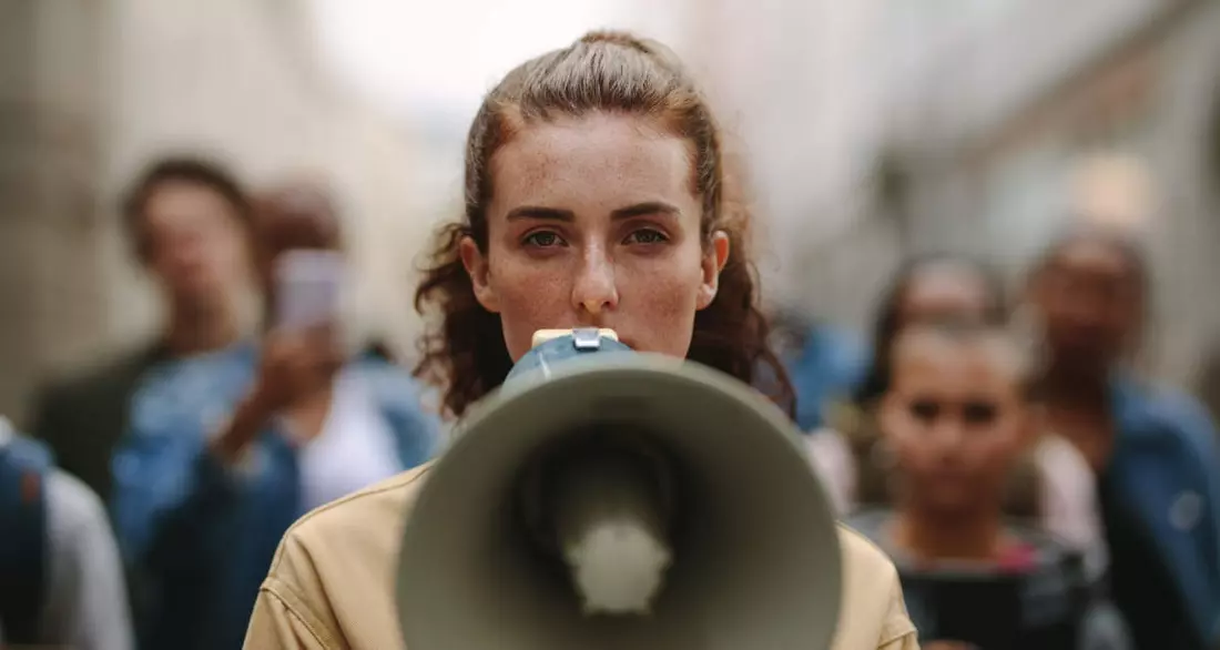 Eine Frau mit einem Megafon, im Hintergrund weitere Menschen. Foto: Adobe Stock